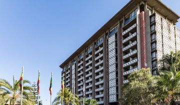 The tall concrete tower of the Hilton Addis Ababa with patterned balconies, palm trees and flags under a blue sky