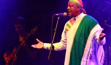 Mahmoud Ahmed in a white robe, a green and white shawl and a colorful head scarf sings at a microphone with open arms, a guitarist behind him, Rudolstadt, Germany, 2010