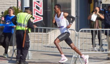 Ethiopian runner Tamirat Tola, in a white vest with a race bib reading TOLA, runs past metal barriers, a police officer and fans on a street in the Bronx during the 2023 New York City Marathon