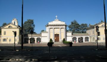 The pale classical entrance of the Monumental Cemetery of Turin, with columns, a cross on top and yellow buildings on both sides, under a clear blue sky in 2013