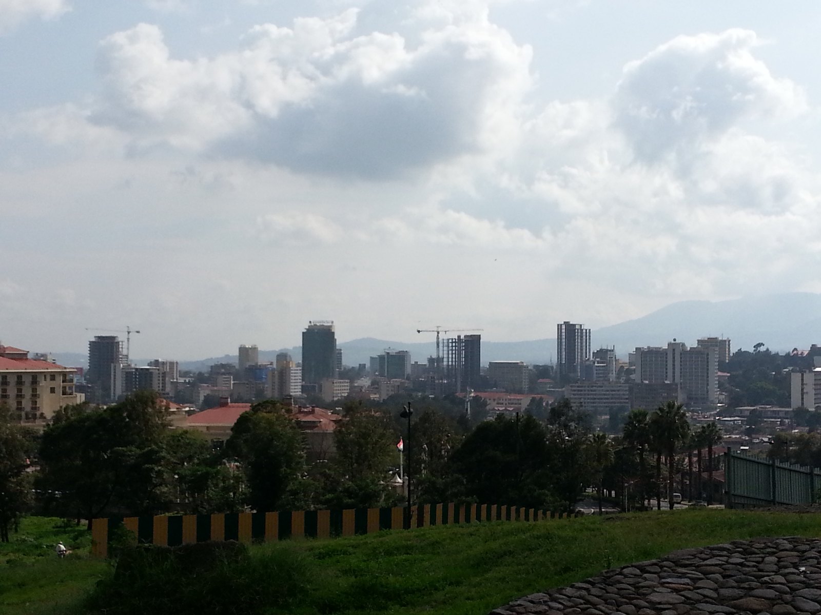 Office towers and cranes rise above trees and red roofs on the Addis Ababa skyline under a cloudy sky, with hills behind, in 2012