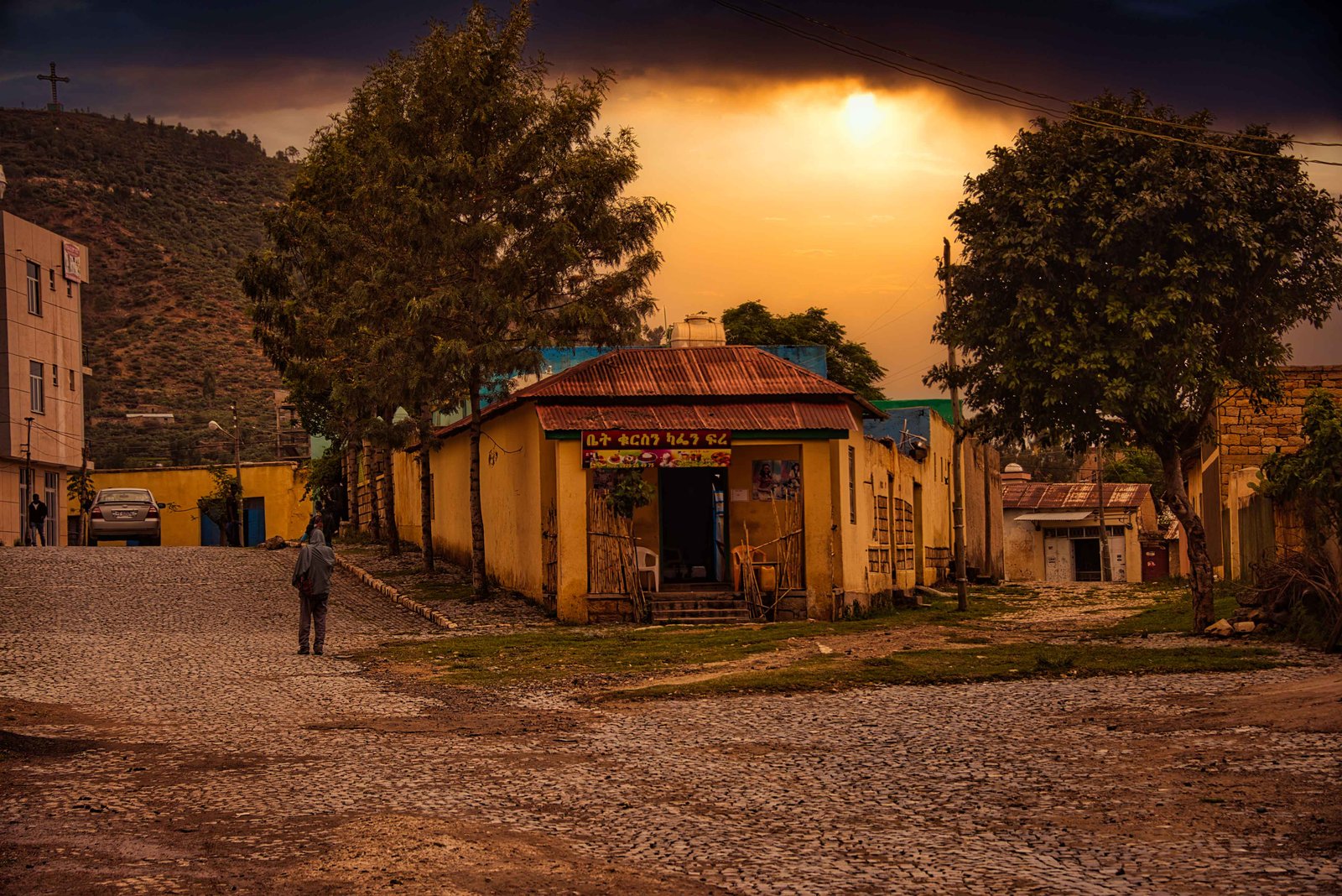 A cobbled street with a small yellow shop and trees under an orange evening sky in Adigrat, Ethiopia, 2019