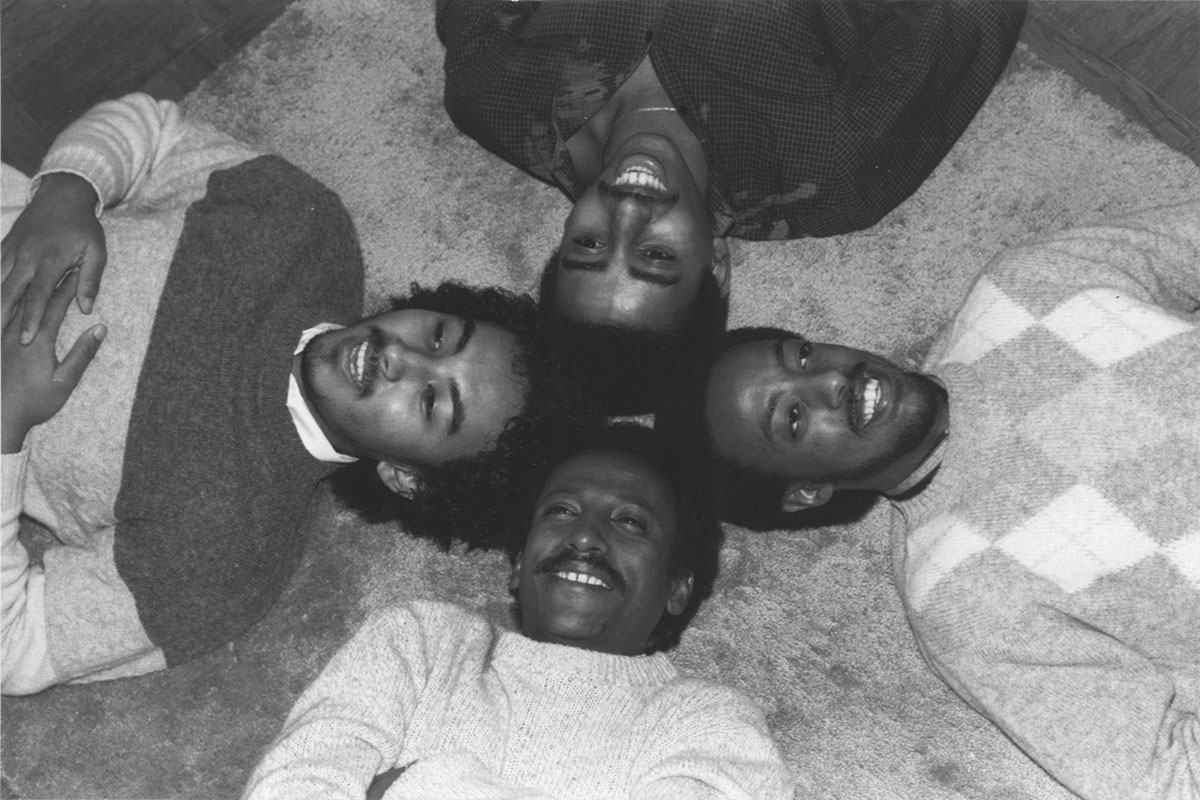 A black and white photo of the four members of Admas lying on a carpet with their heads together, smiling up at the camera, in the 1980s