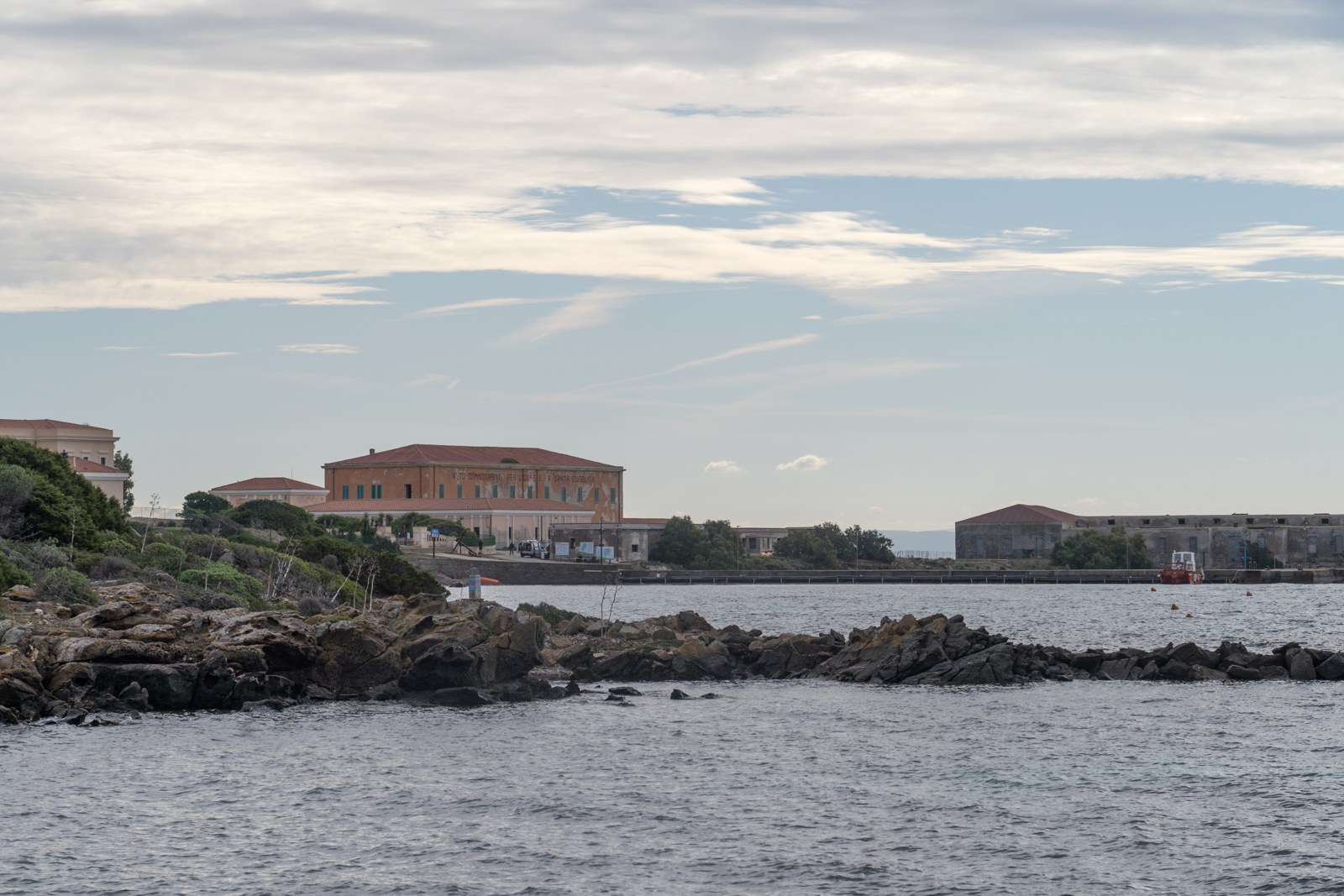 Old red-roofed buildings stand beside a quiet bay with dark rocks in the water at Cala Reale on the island of Asinara, Sardinia, in 2024