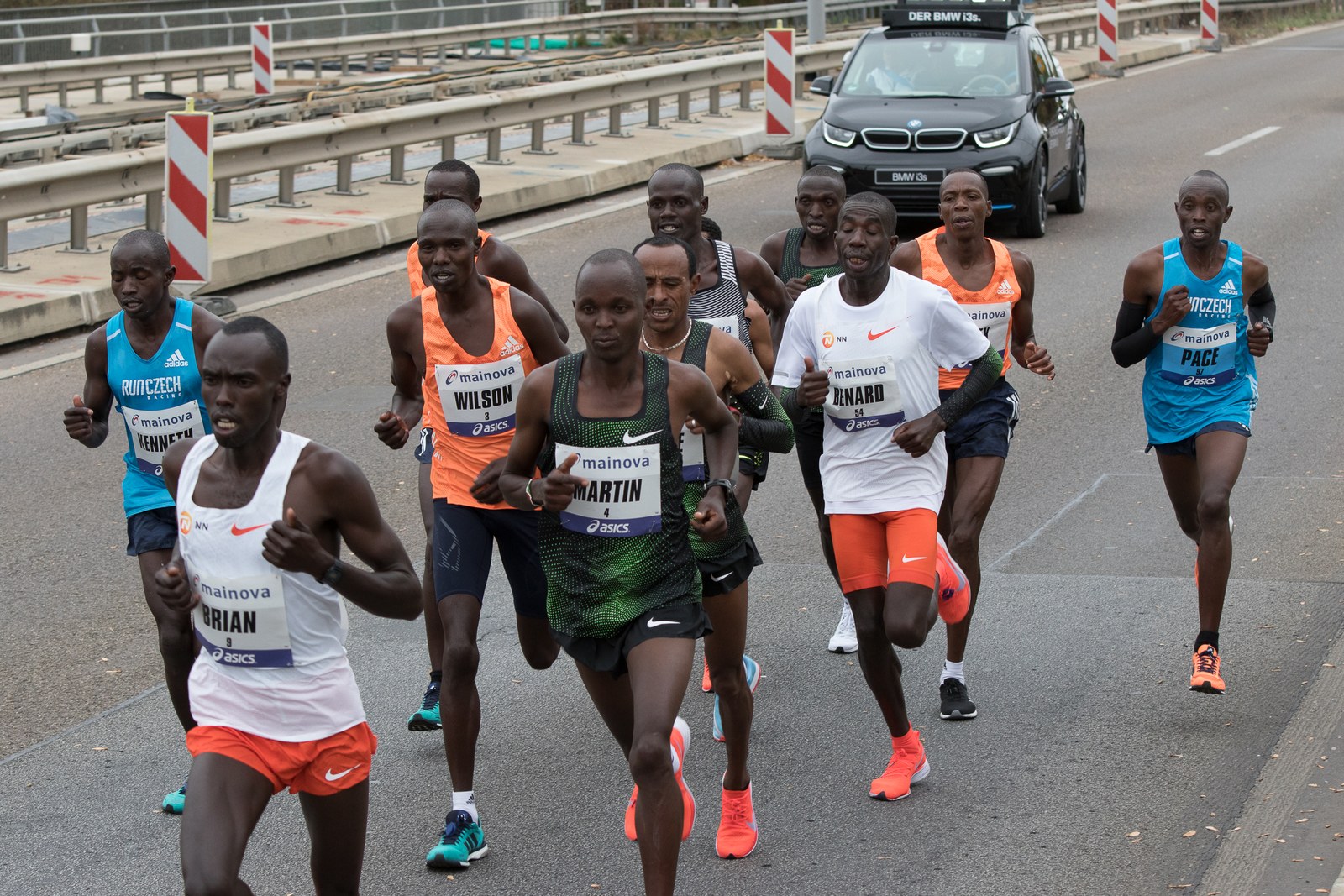 A group of male elite runners, several wearing Mainova race bibs and two pacemakers, run on a wide road at the 2018 Frankfurt Marathon