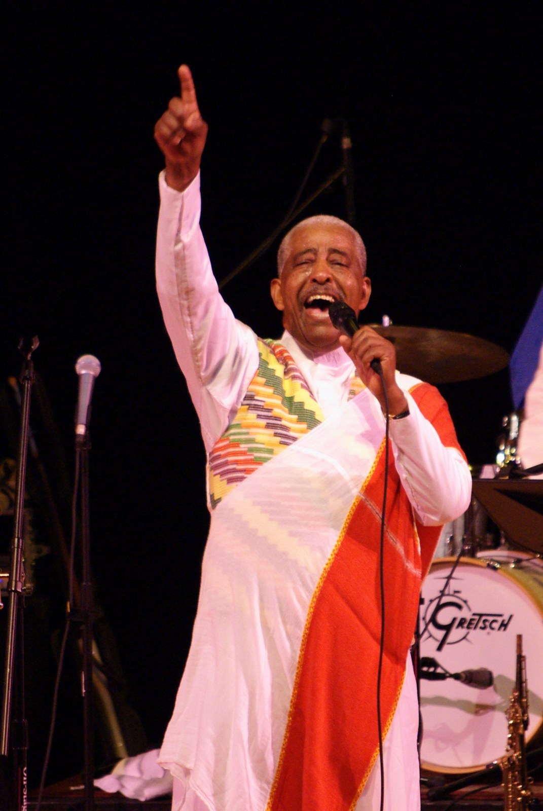 Mahmoud Ahmed in white traditional clothes and a woven shawl raises one hand as he sings into a microphone on a dark stage in Denver, 2009
