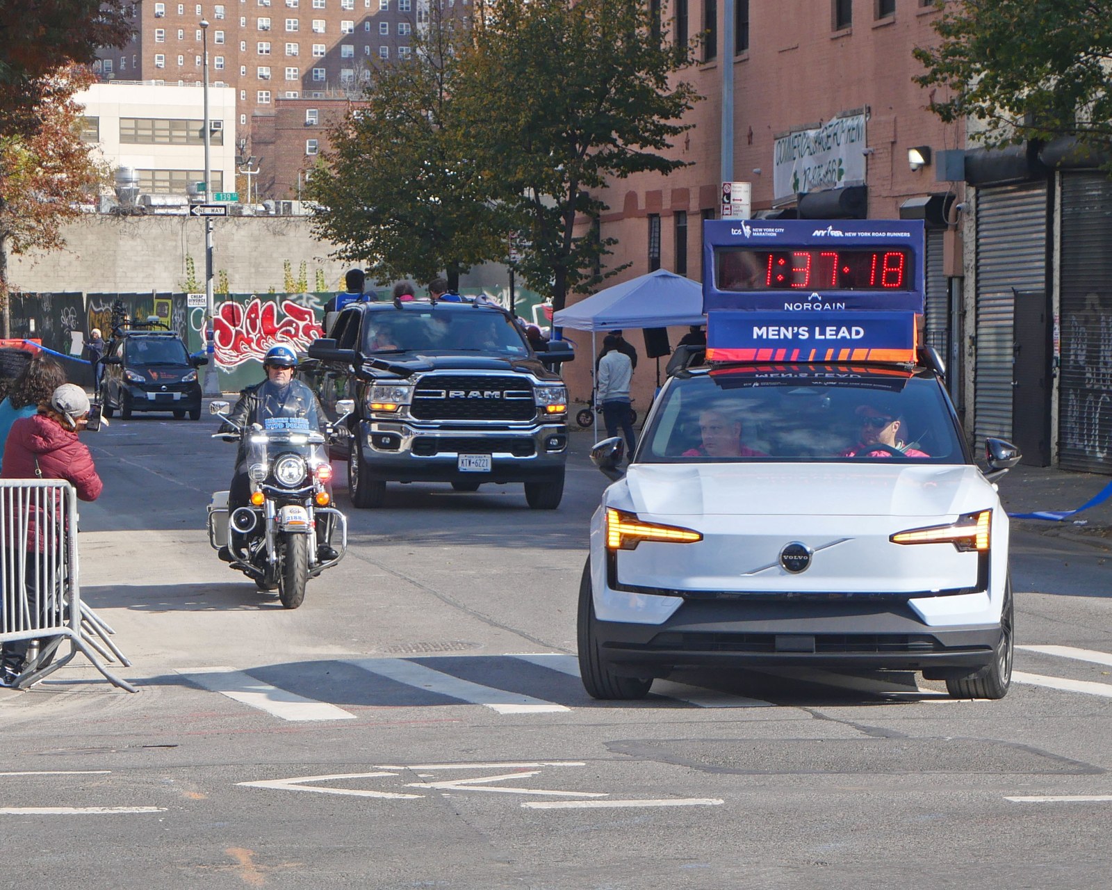 A white car with a sign reading Men's Lead and a red race clock on its roof drives down a Bronx street, followed by a police motorcycle and a black pickup truck, during the 2023 New York City Marathon