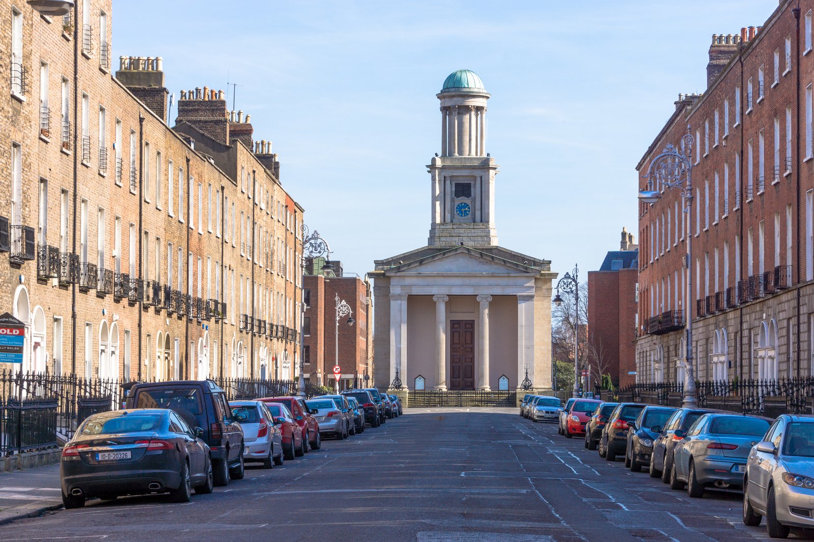 A quiet Dublin street with brick houses and parked cars on both sides, leading to St Stephen's Church, a pale church with columns and a domed tower, under a blue sky in 2015
