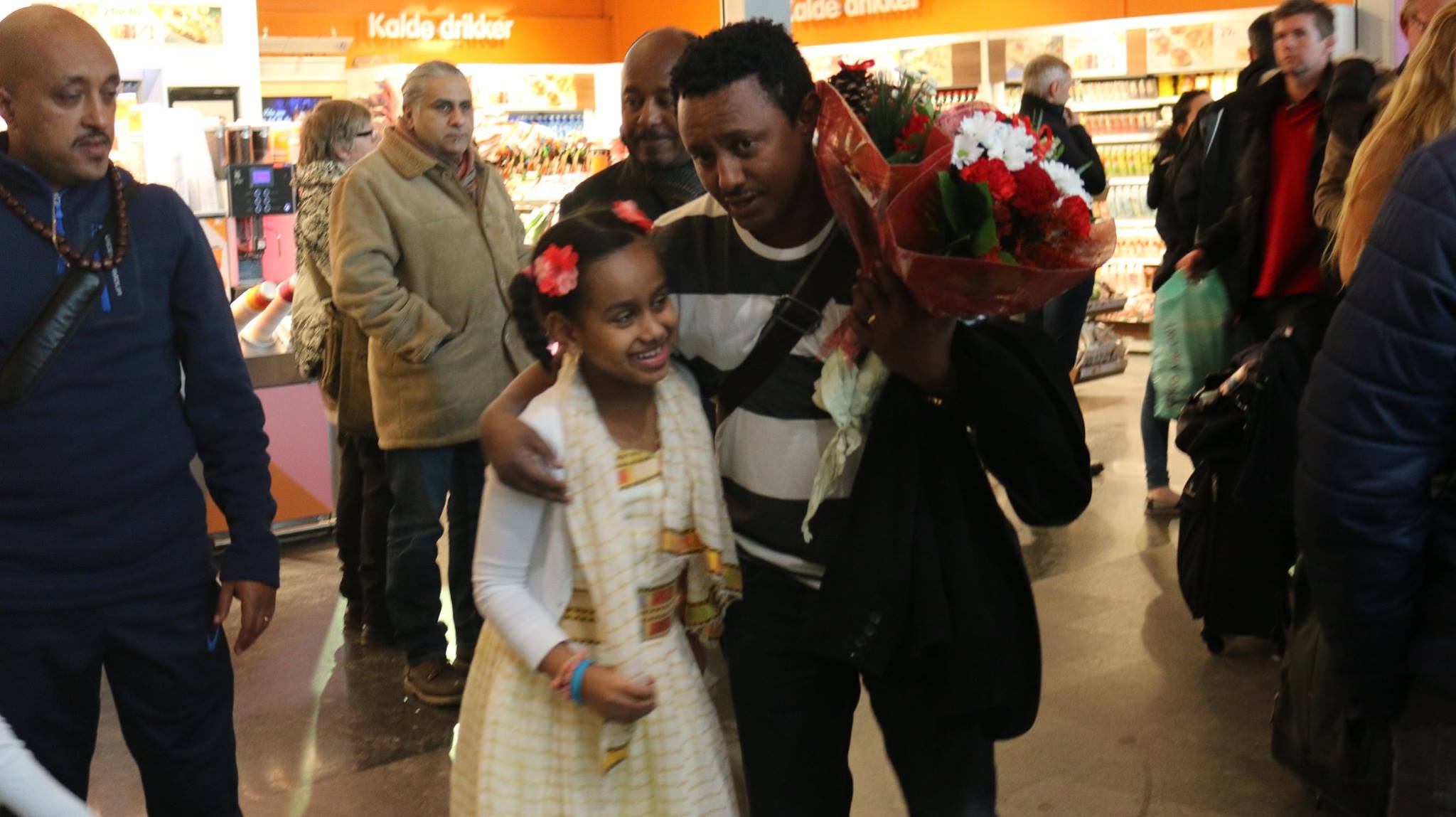 Teddy Afro, holding flowers, smiles with a young girl in traditional Ethiopian dress as fans greet him at Oslo Airport in 2014